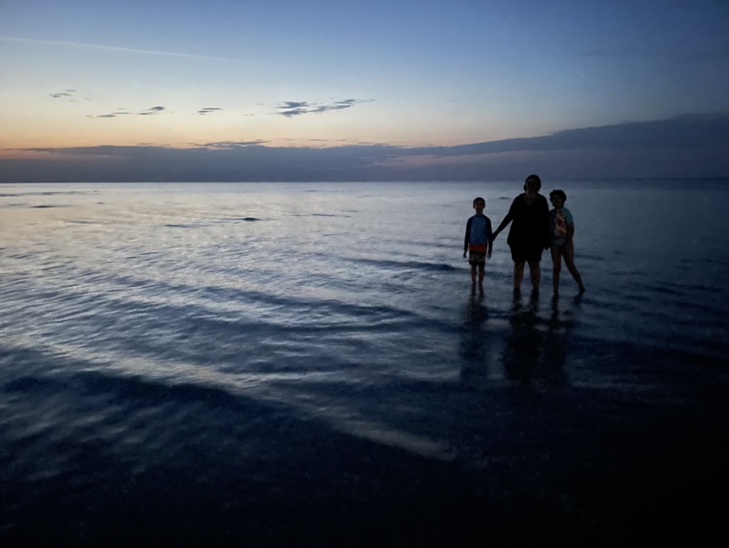 A photo of my family taken at Cedar Island, NC in 2022: An adult and two children standing in ankle-deep water at the beach, in silhouette against a darkening sunset sky.